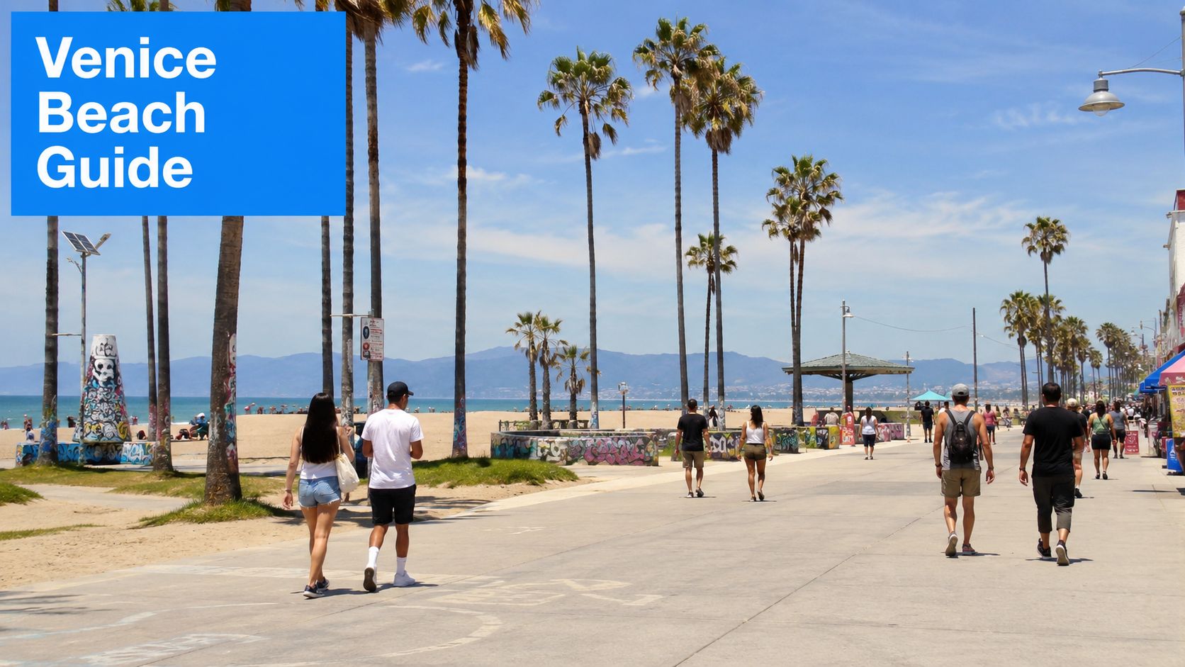 People strolling along the sunny Venice Beach boardwalk with palm trees and a sandy beach in view.